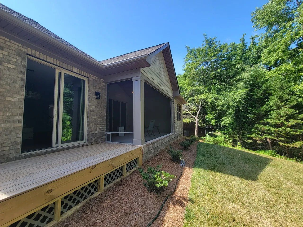 Front porch of a house with screened windows, wood siding, and a grassy yard with trees.