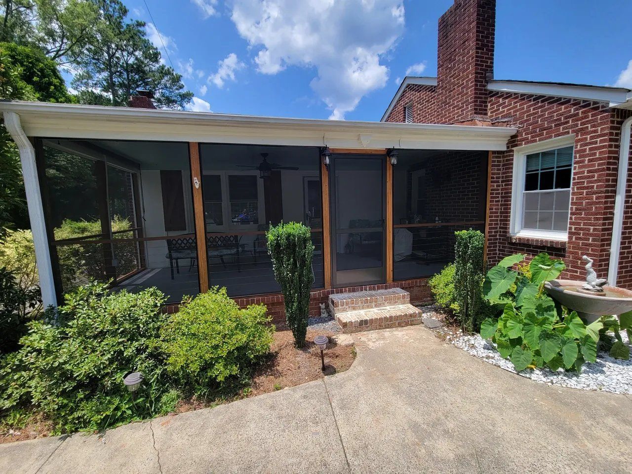 Brick house screened porch with white roof, concrete patio, and green shrubs under a blue sky