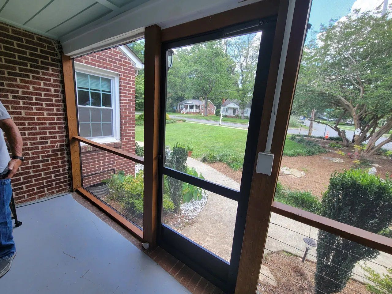 Covered porch with screen door and brick wall overlooking a green yard and trees