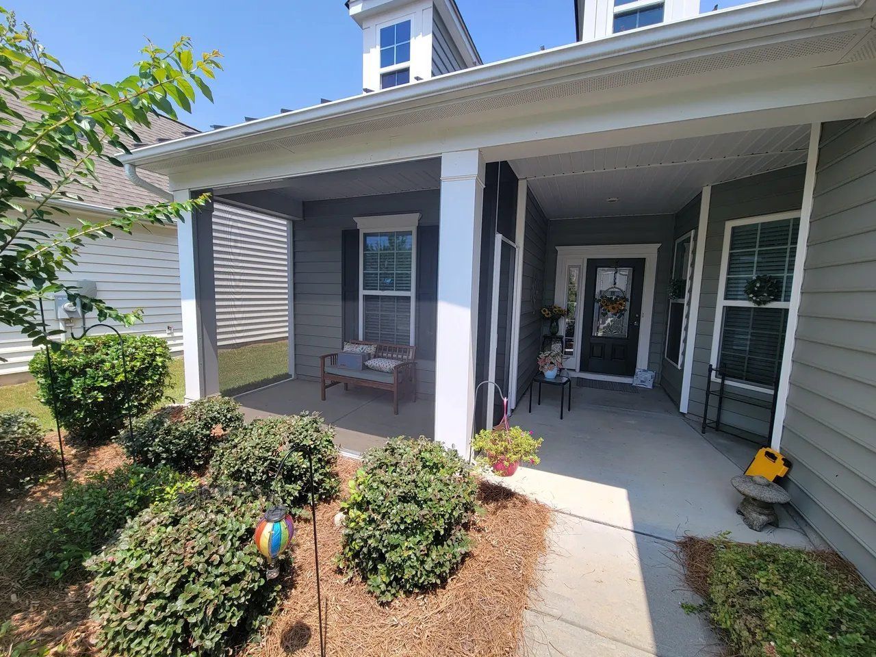 Front porch of a house with white columns, gray siding, potted flowers, and a small chair by the entrance.