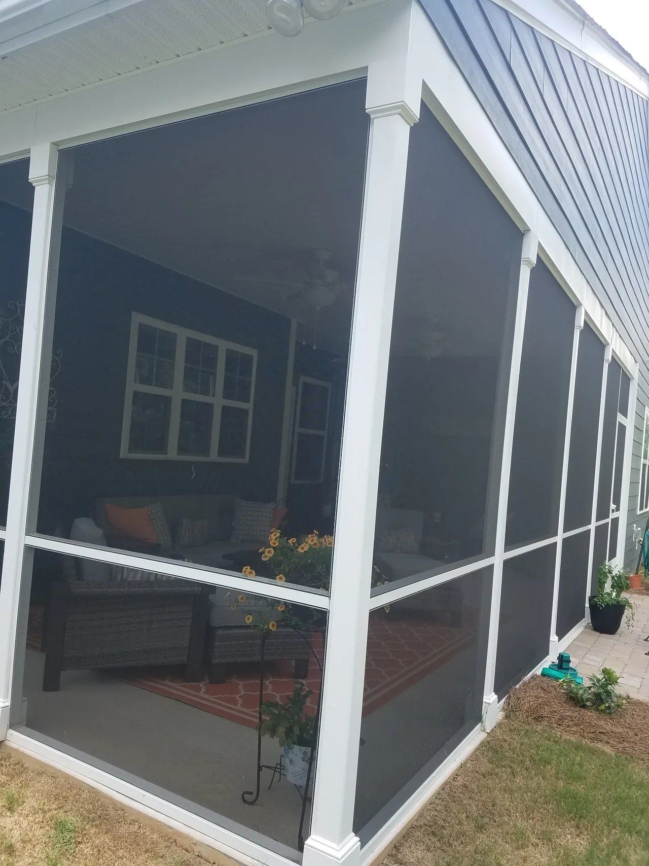 Screened porch with white columns, dark furniture, and potted plants on a brick floor beside a house.