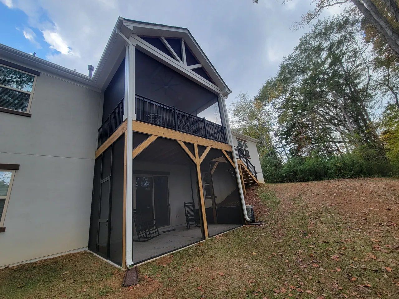 Two-story house exterior with a covered balcony, wooden supports, and a sloped roof beside a grassy slope.