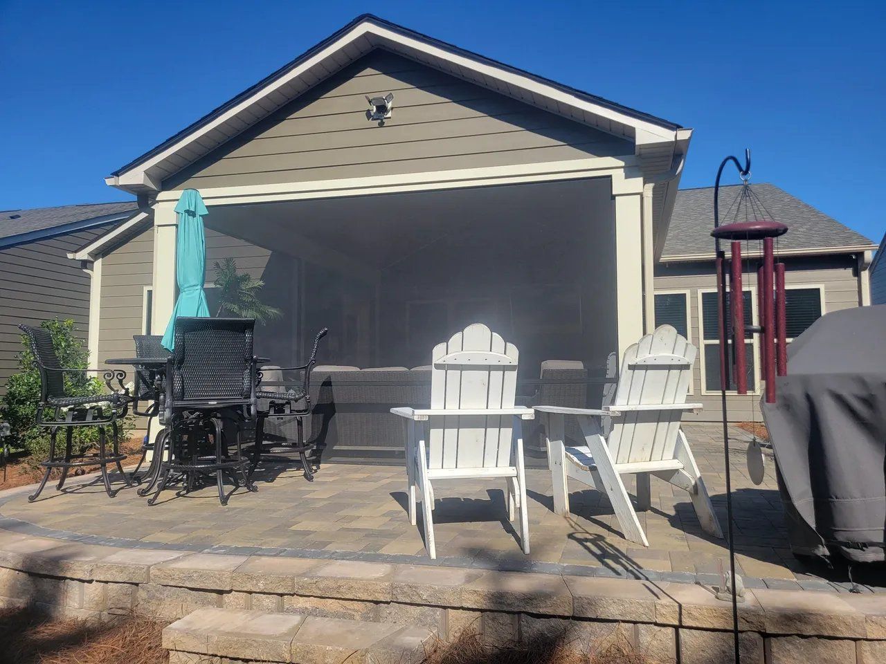 Patio with white chairs, table, grill, and a turquoise umbrella in front of a house.
