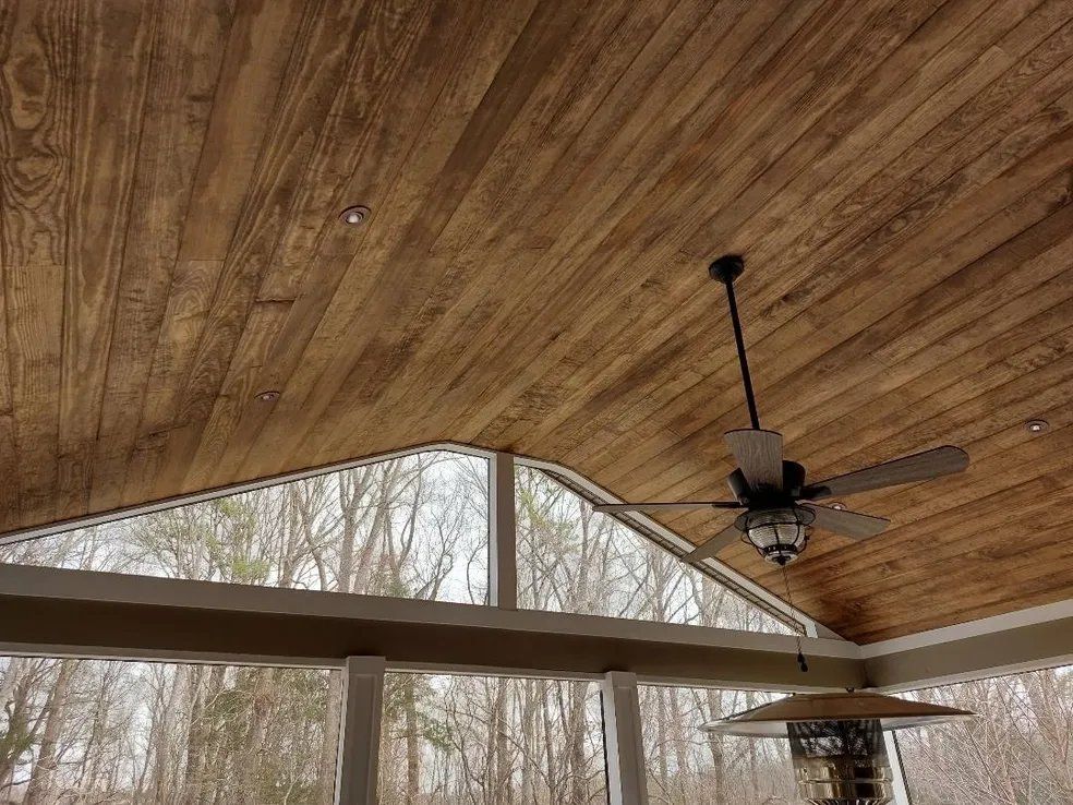Covered patio with wood-paneled ceiling, ceiling fans, and large windows overlooking trees