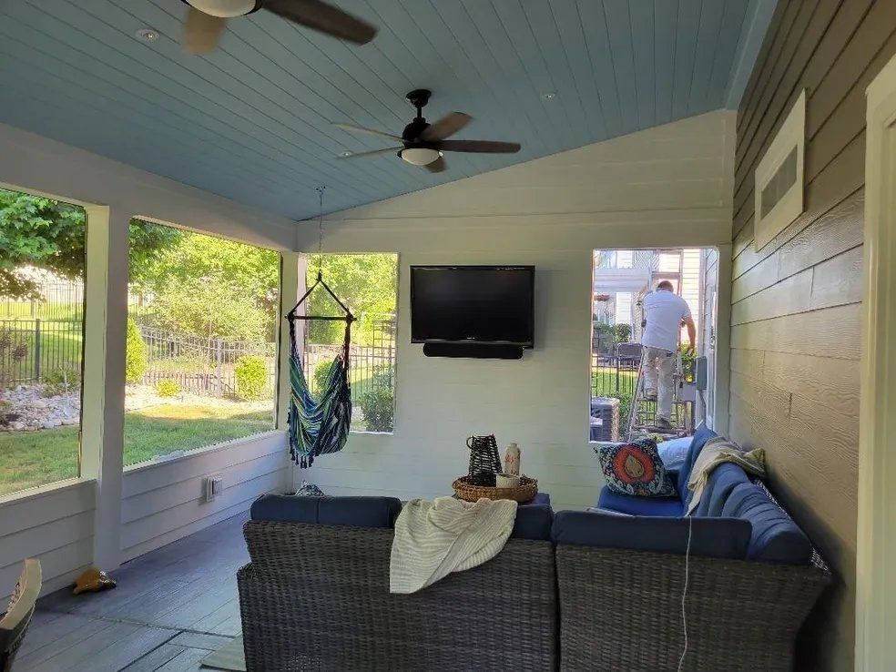 Covered porch with dark wicker sofa, hanging chair, wall TV, ceiling fans, and views of a backyard garden