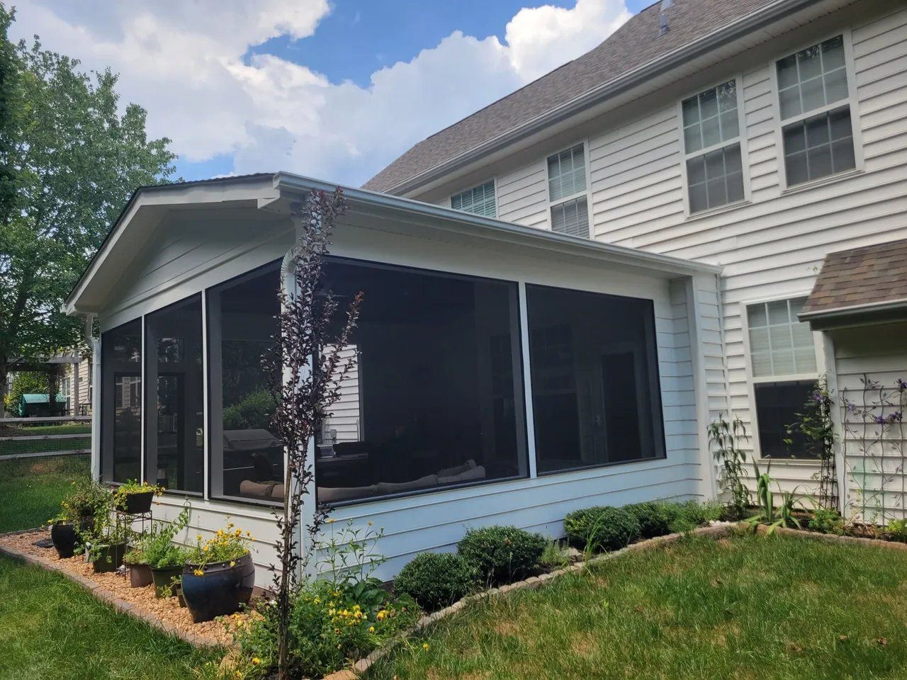 Screened-in sunroom with black mesh panels attached to a white house and small landscaped garden.