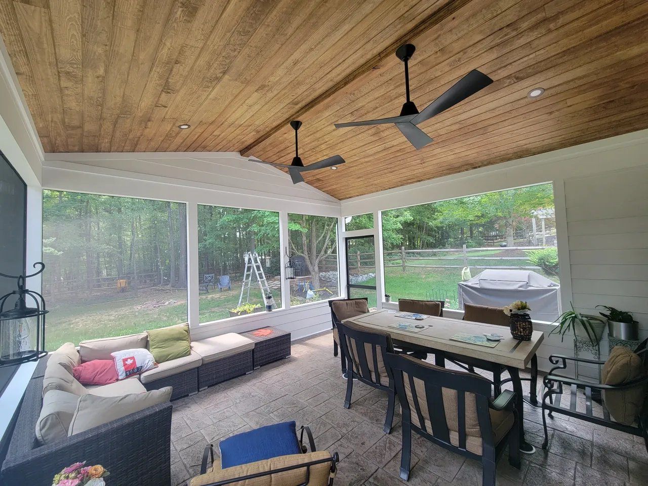 Covered outdoor patio with wicker seating, dining table, ceiling fans, and screened view of a backyard and grill.