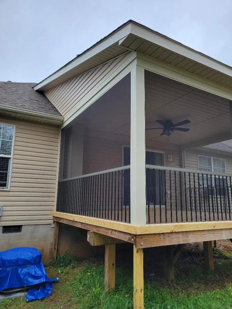 Small screened porch on a house with a black railing and a ceiling fan; blue tarp on the grass nearby.