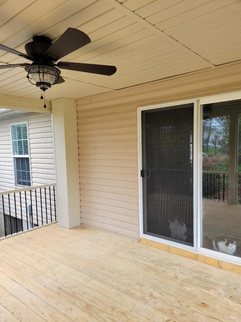 Covered porch with black ceiling fan, beige siding, and screened sliding door overlooking a railing