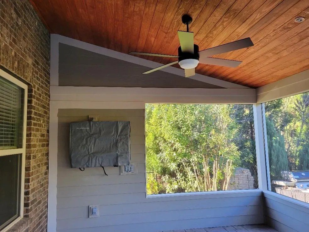 Covered porch with wood ceiling, ceiling fan, gray siding, and view of trees through a large opening