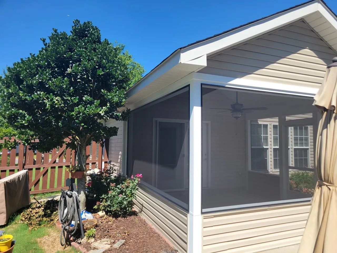 Small beige shed with screened porch and white trim in a backyard, with a bicycle and flowers nearby.