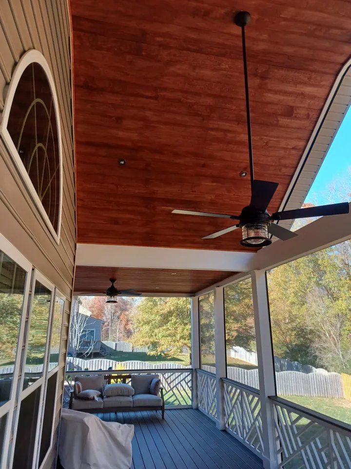 Covered porch with wood ceiling, ceiling fan, sofa, and railing overlooking a yard and trees.