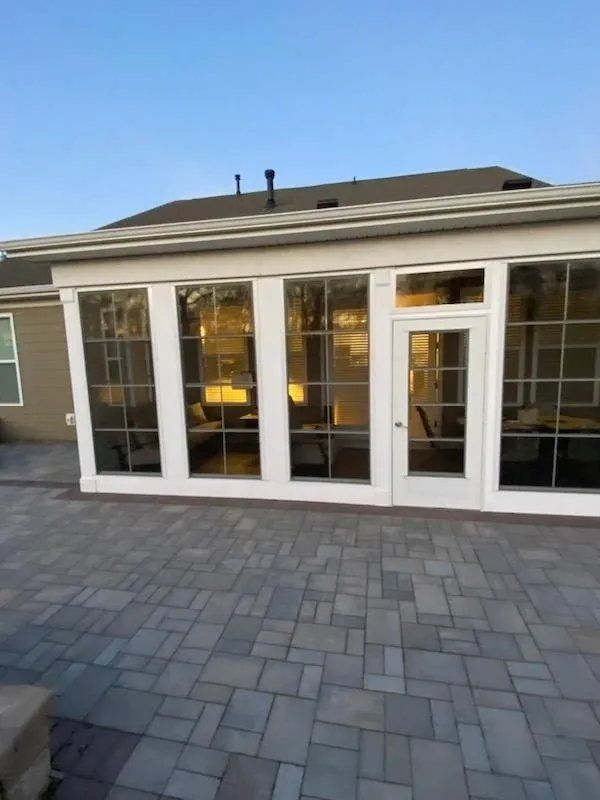 Glass-enclosed patio with white-framed doors and windows beside a paved stone terrace