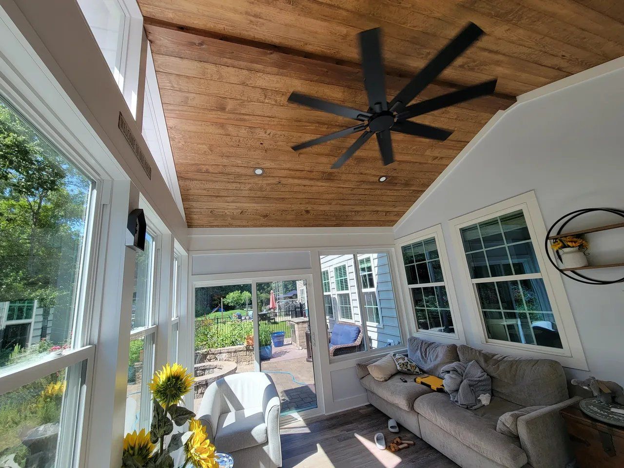 Screened porch with gray sofa, white chairs, wood ceiling, ceiling fan, and garden view