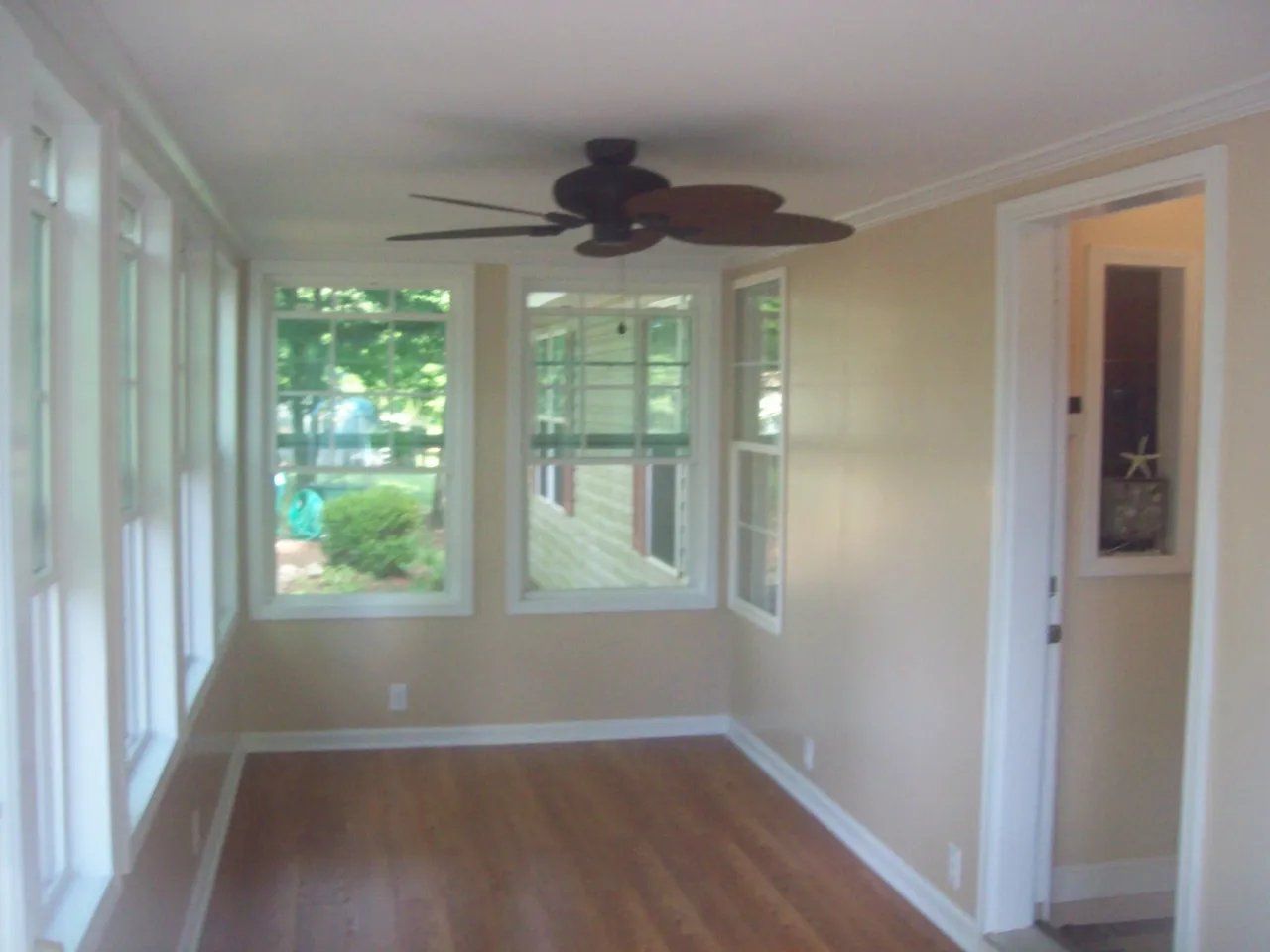 Bright empty sunroom with wood floor, white trim, ceiling fan, and large windows overlooking greenery