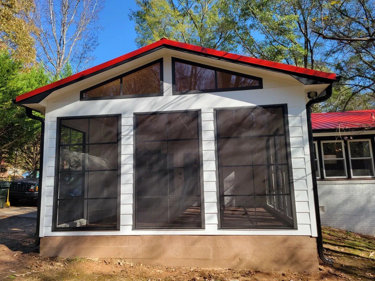 Small white building with red roof trim and three screened windows in a wooded outdoor setting