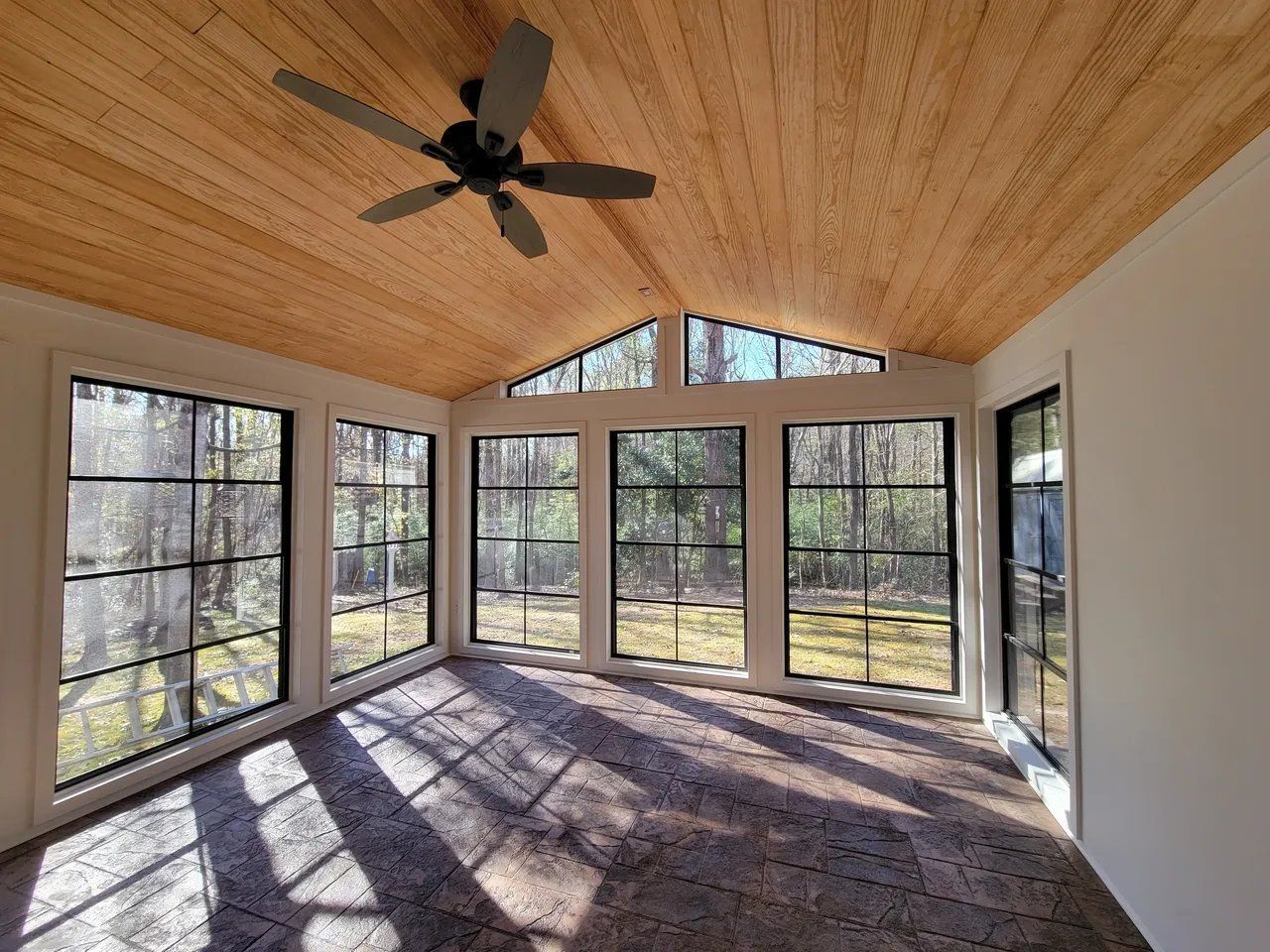 Sunlit screened porch with wood ceiling, ceiling fan, and large windows overlooking trees