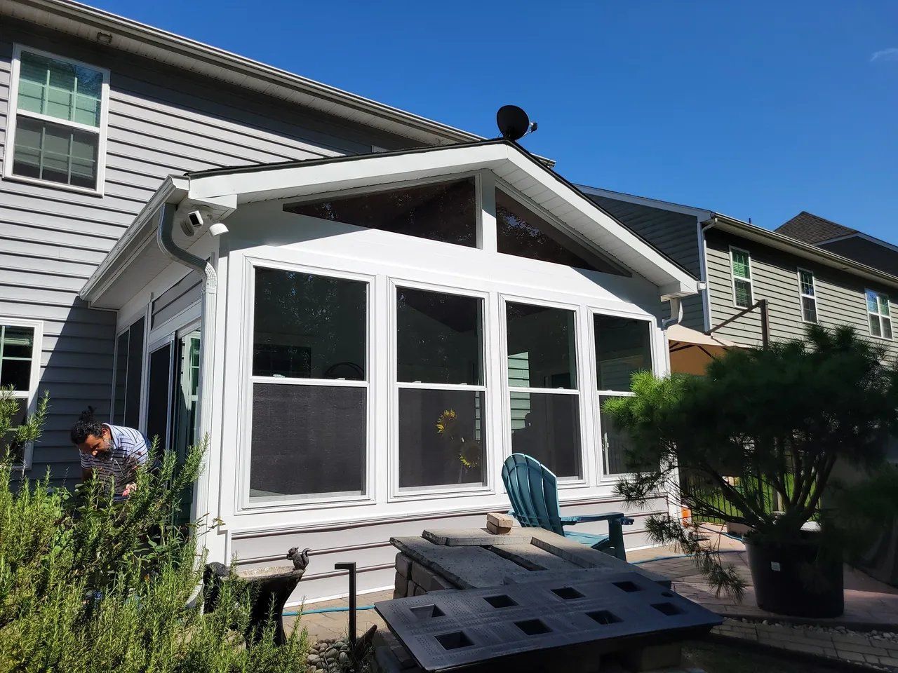 White sunroom extension with large windows on a house patio, beside a deck and shrubs under a blue sky