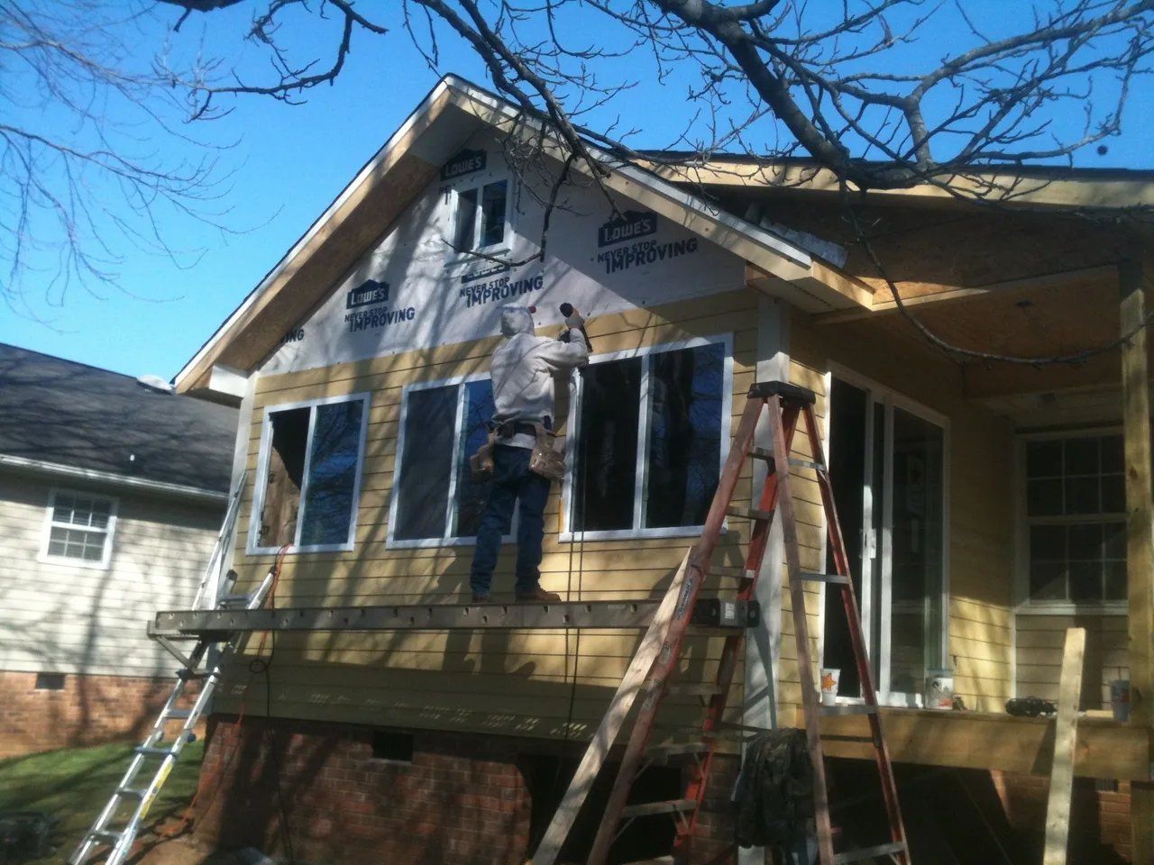 Two workers on ladders painting the front of a yellow house under a bright blue sky
