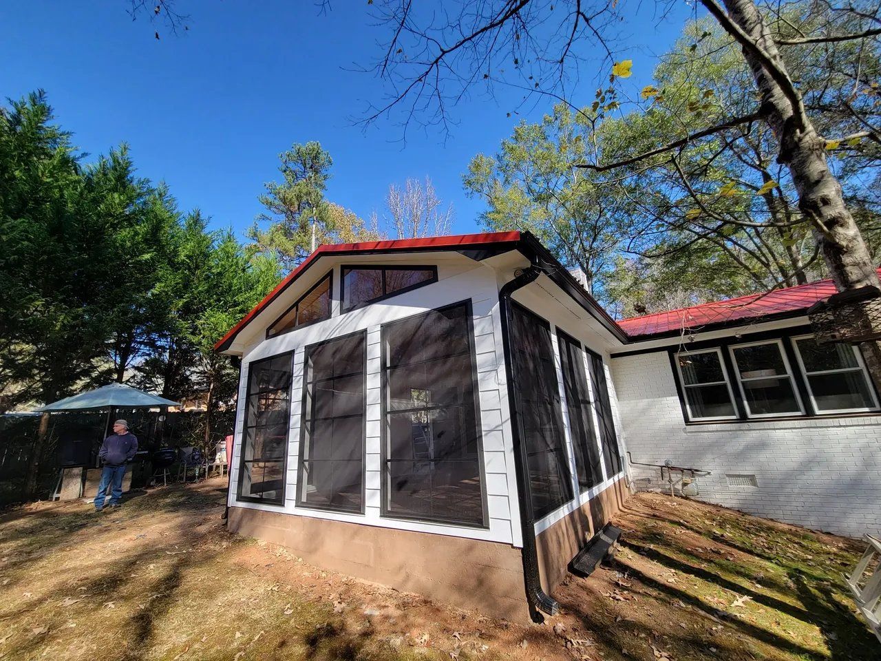 Screened porch addition with red roof beside a house, surrounded by trees and a blue sky.