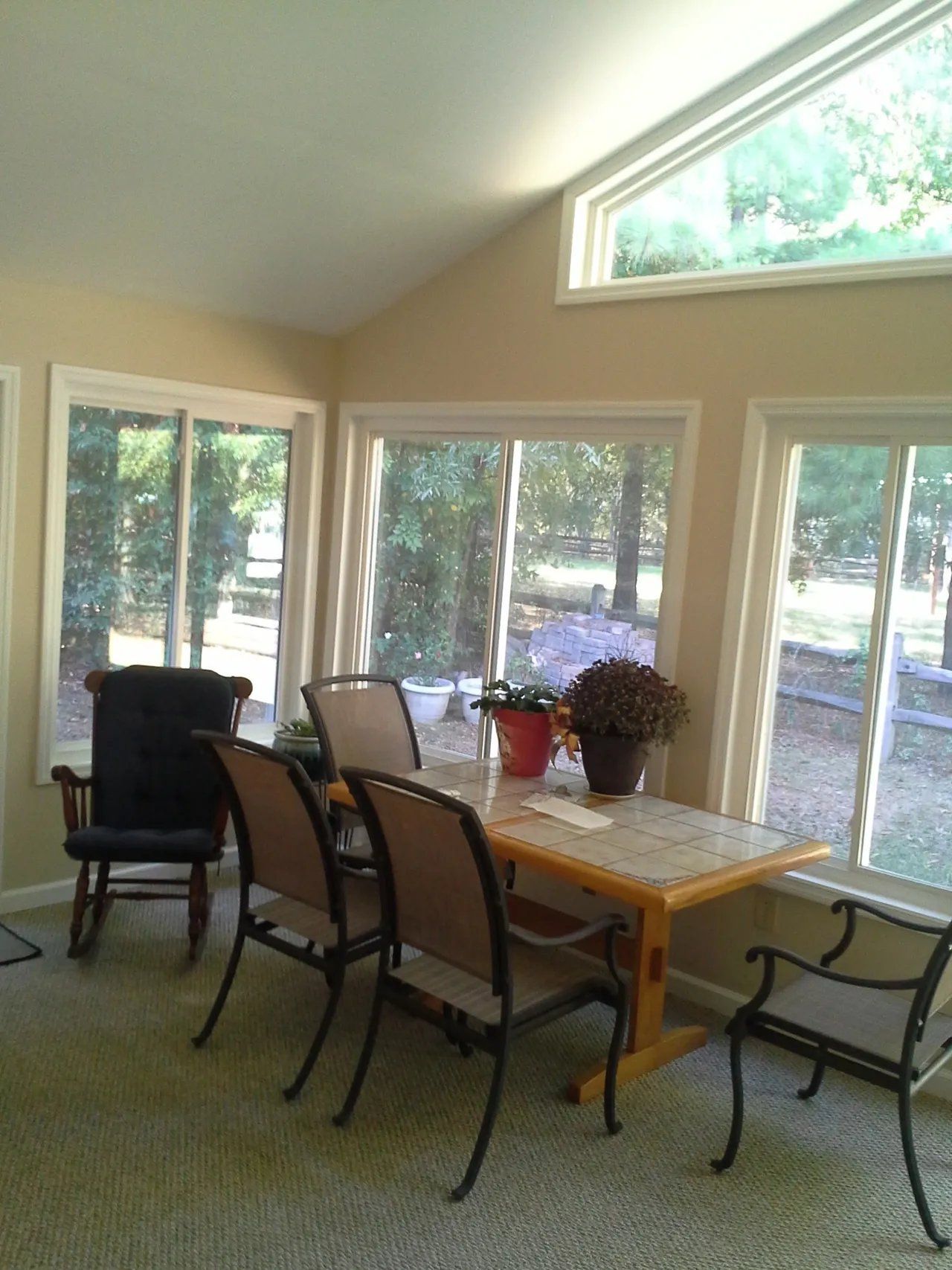 Sunlit dining room with wooden table, chairs, and large windows overlooking trees