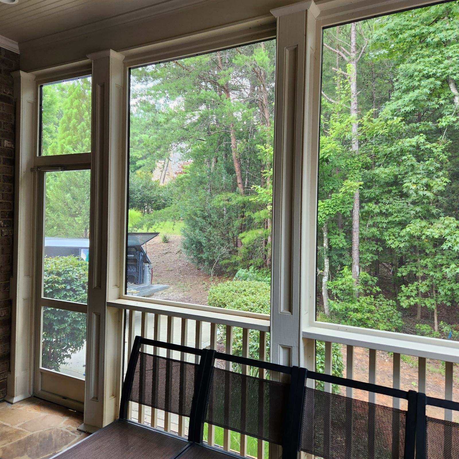 Covered porch with white railings and black chairs overlooking a green wooded yard and driveway