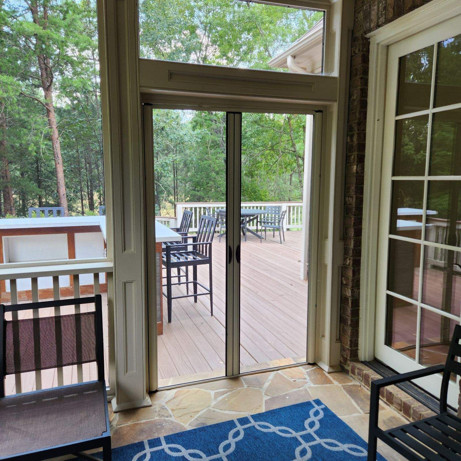 Interior view of a screened porch with open French doors, wood deck, chairs, and trees outside
