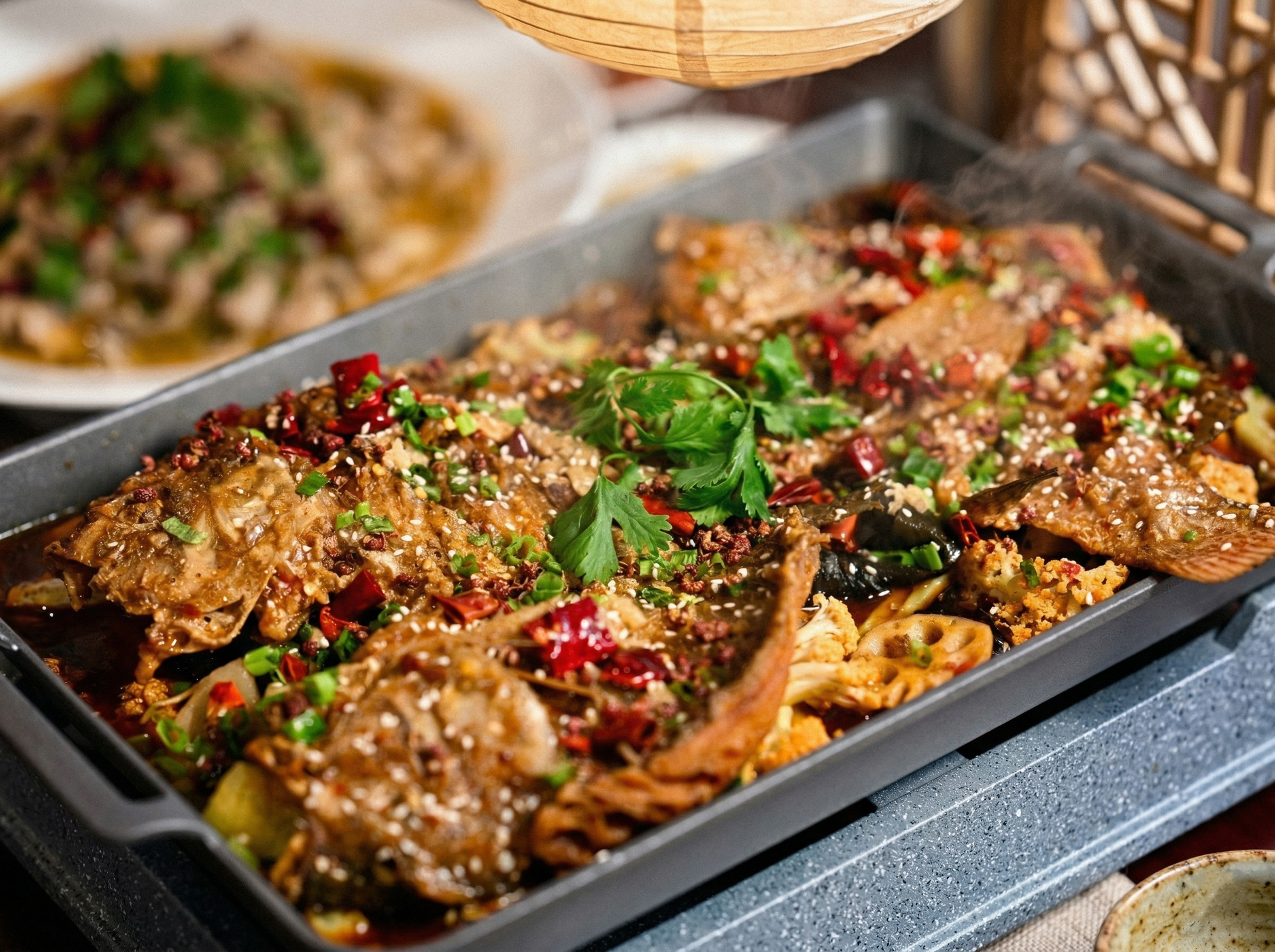A sizzling rectangular pan of spicy braised whole fish topped with cilantro, sesame seeds, and peppers.