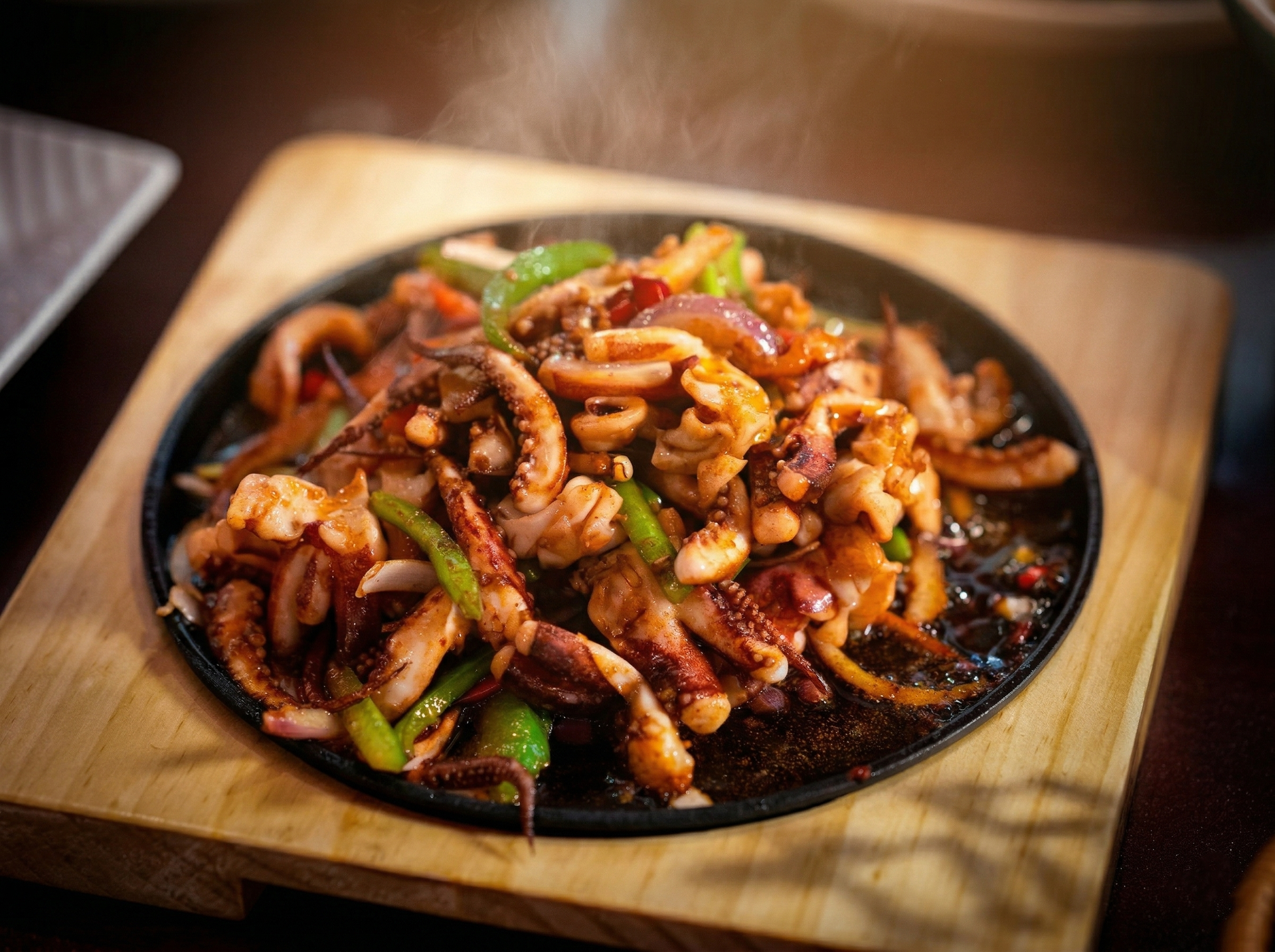 A hot, steaming skillet of stir-fried squid with green bell peppers and onions served on a wooden board.
