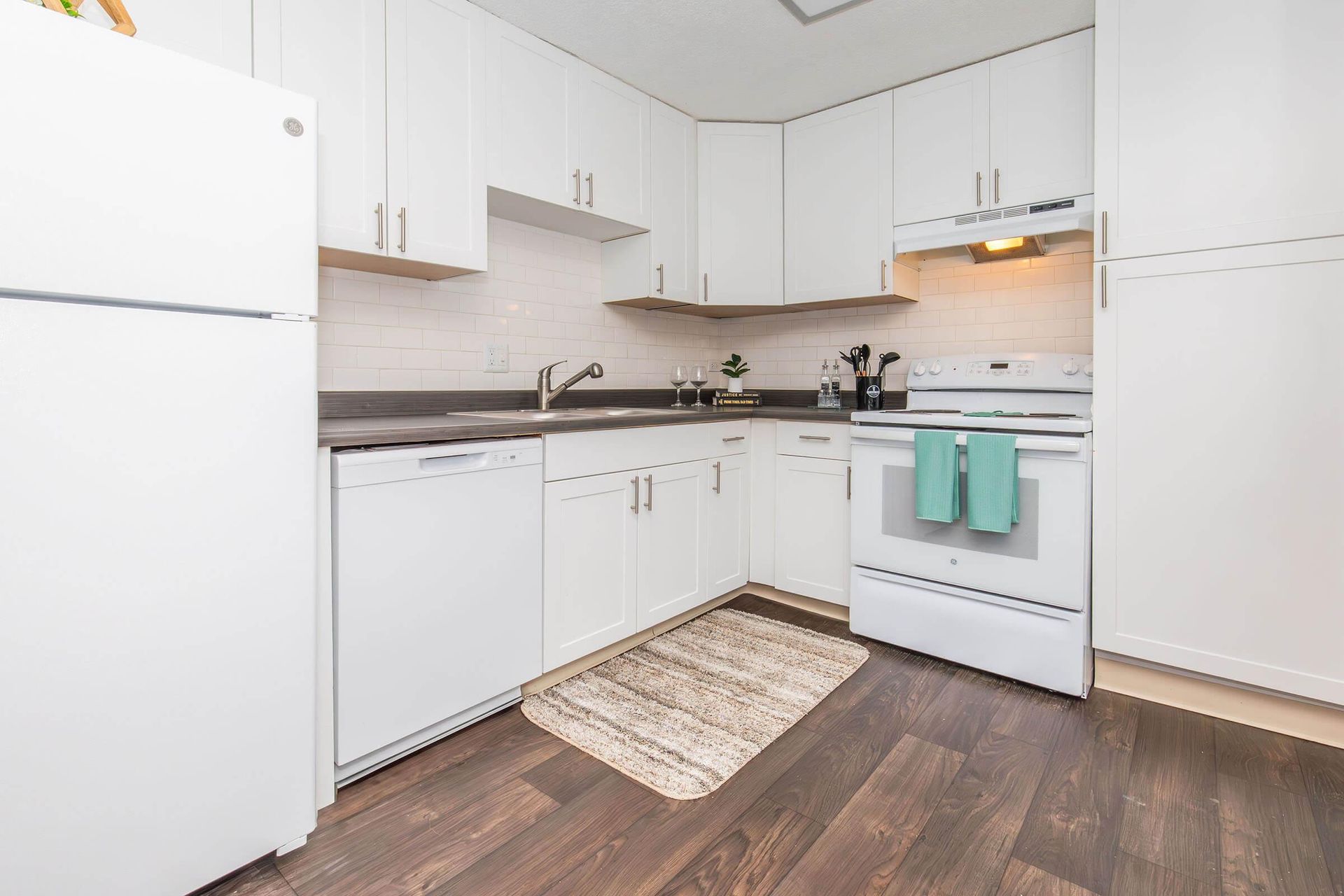 White kitchen with white cabinets, appliances, dark wood floors and small rug.