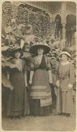 Four women in hats and long dresses pose outdoors in front of foliage and a gazebo.