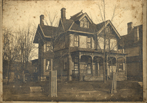 Two-story house with gabled roof, porch, and tall windows in a vintage photo. Bare trees and a street are in the foreground.