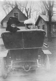 Two women in large hats in a vintage car, buildings in background.
