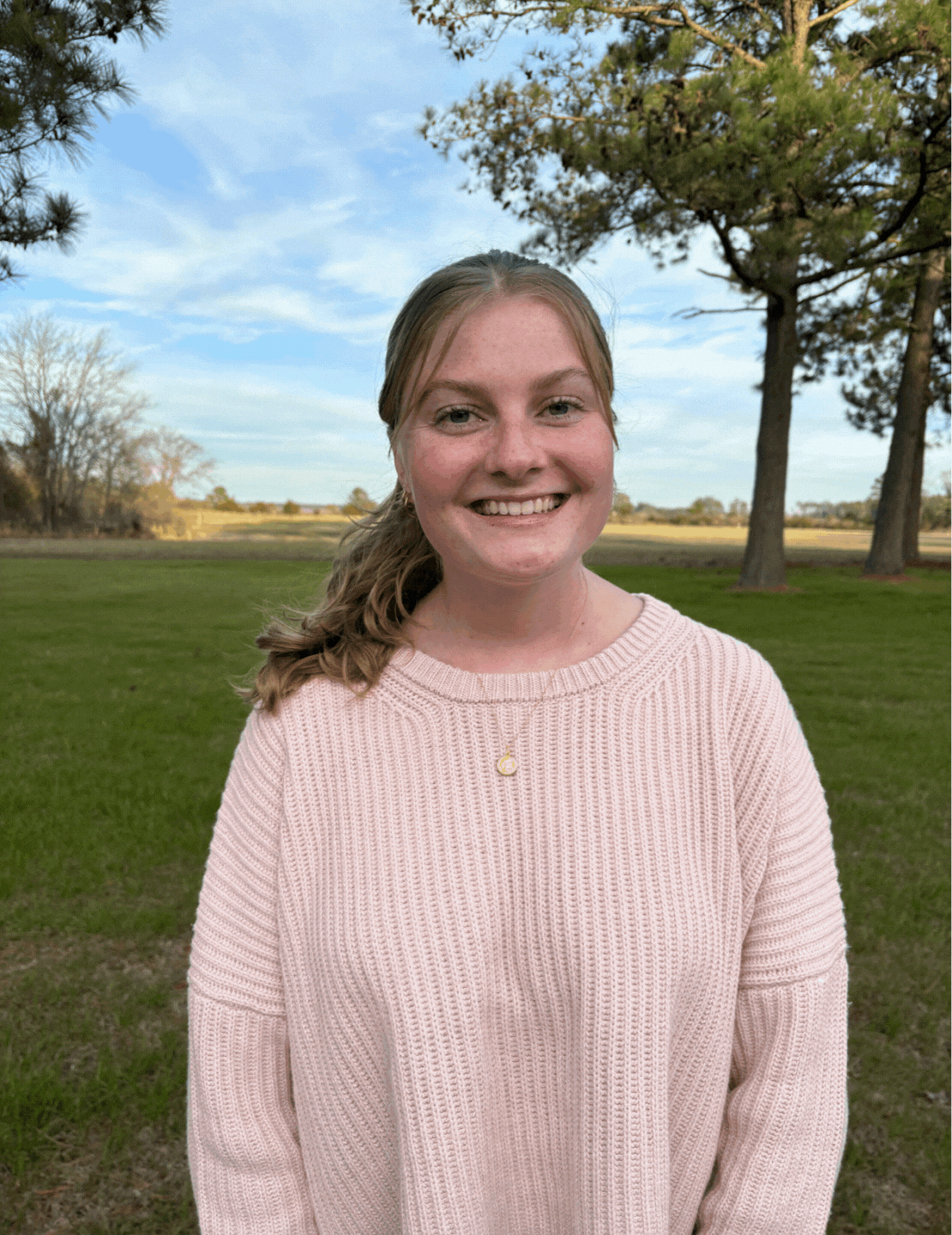 A woman in a pink sweater is standing in a field with trees in the background.