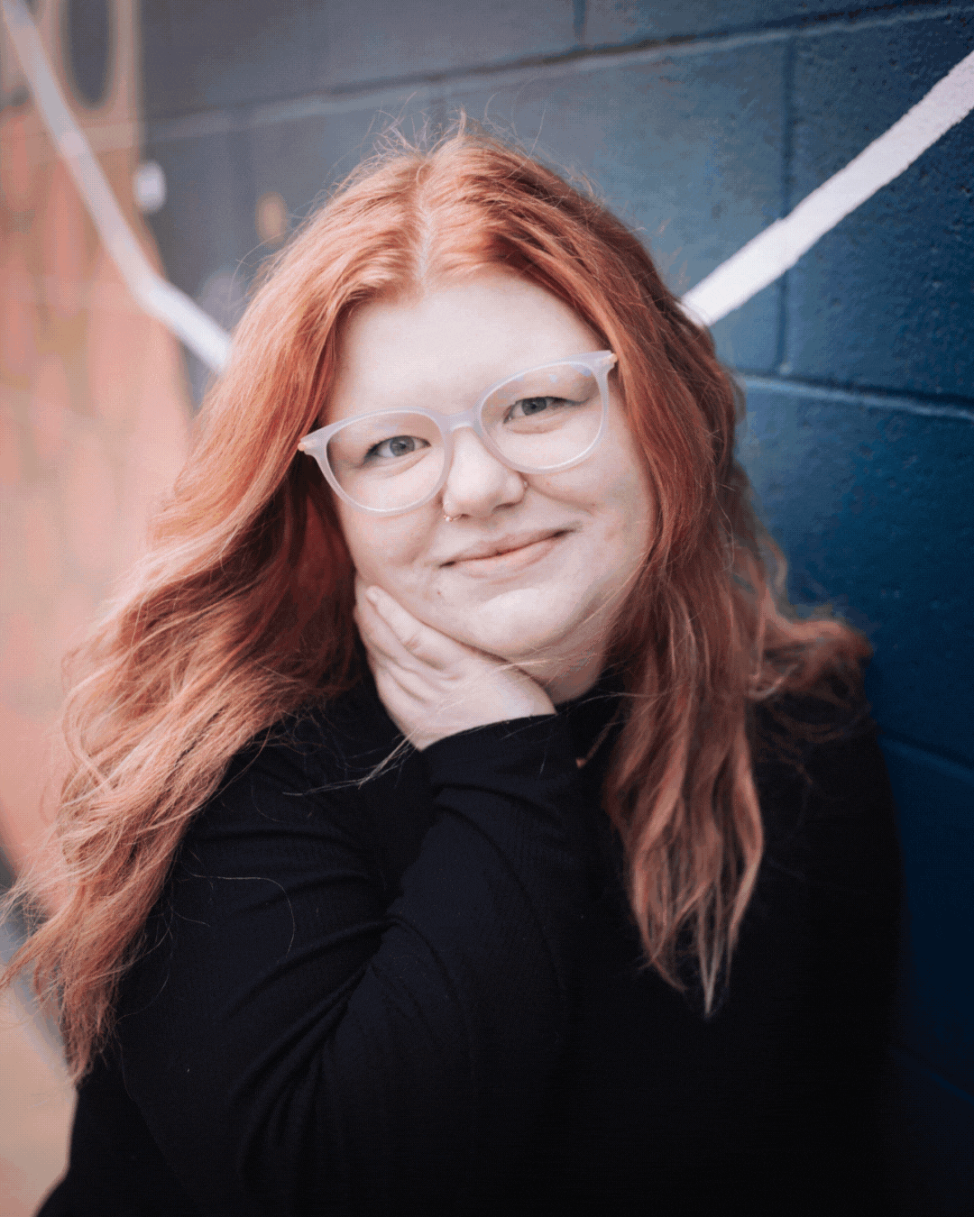 A woman with red hair and glasses is standing in front of a blue brick wall.