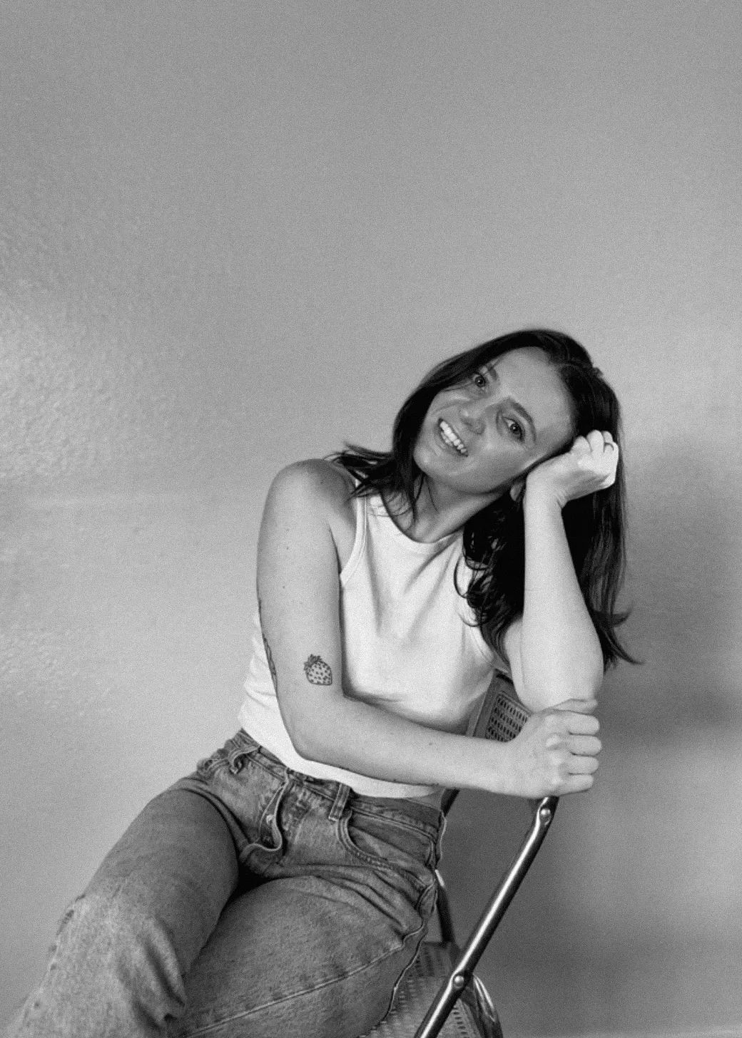 Black-and-white portrait of a smiling person seated on a chair, resting their head on one hand.