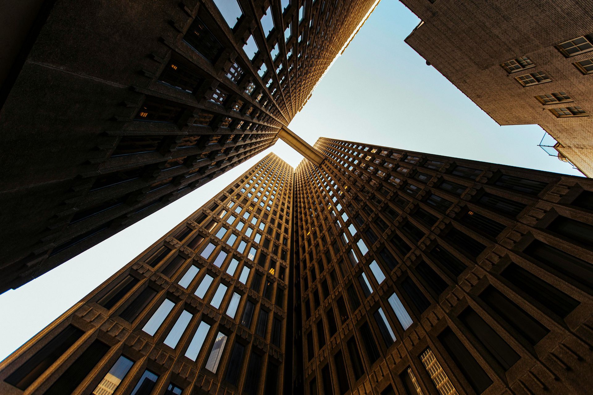 Low-angle view of two tall brick buildings against a blue sky, connected by a walkway.