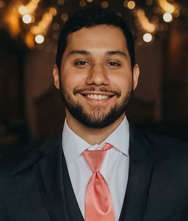 Smiling man in a dark suit with a pink tie against a warm, blurred background