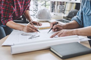 Two people review architectural plans at a desk with a hard hat and rolled blueprints nearby.