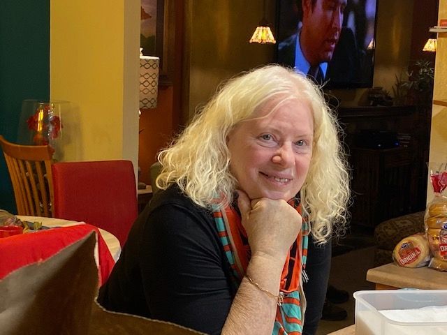 A woman with blonde hair is sitting at a table with her hand on her chin.