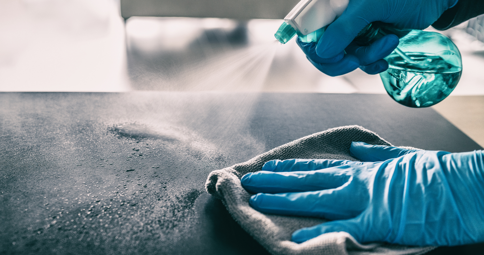 A person wearing blue gloves is cleaning a table with a cloth and spray bottle.