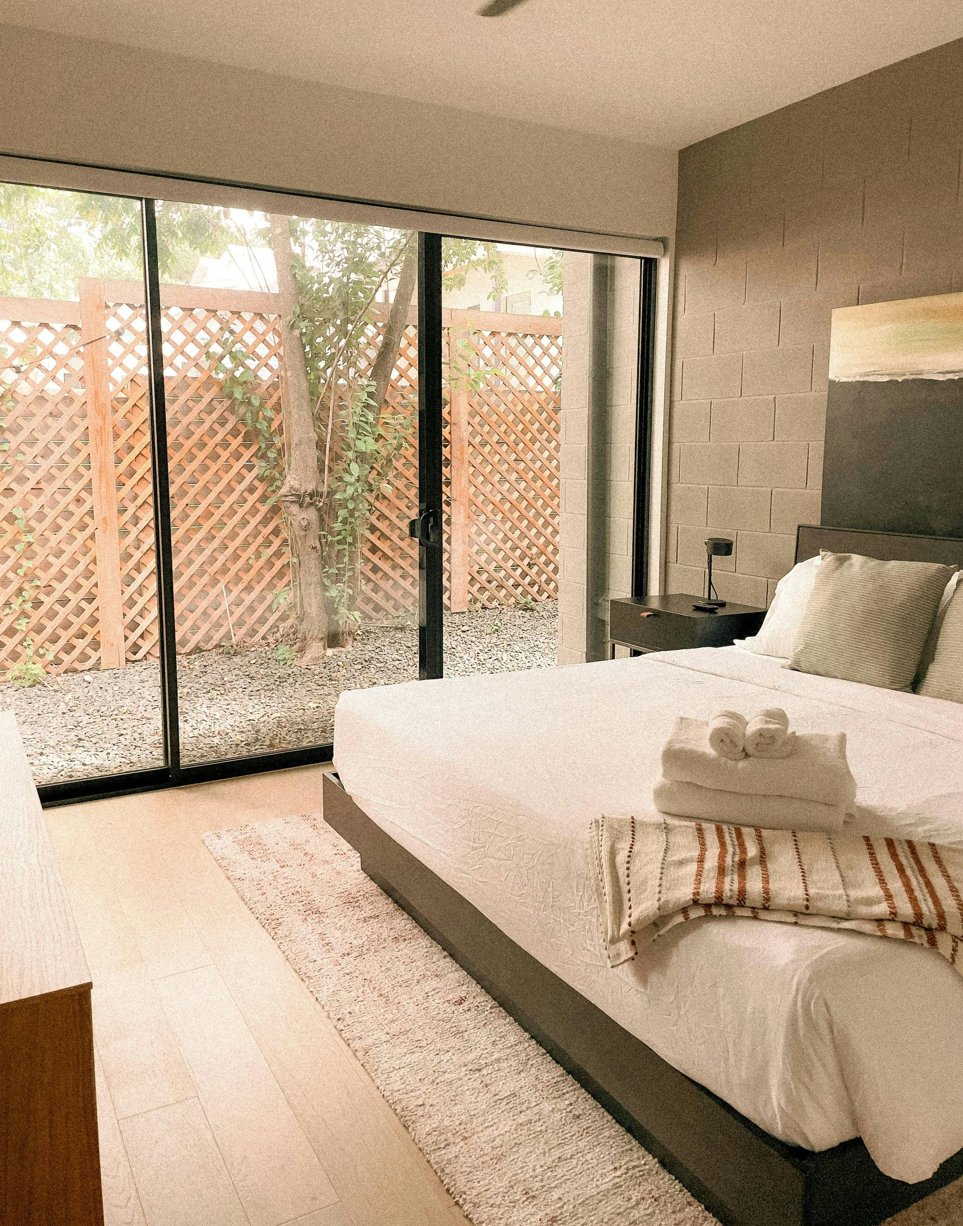 A hotel bedroom with a large bed, light wood floors, a patterned rug, and floor-to-ceiling glass doors opening to a yard.