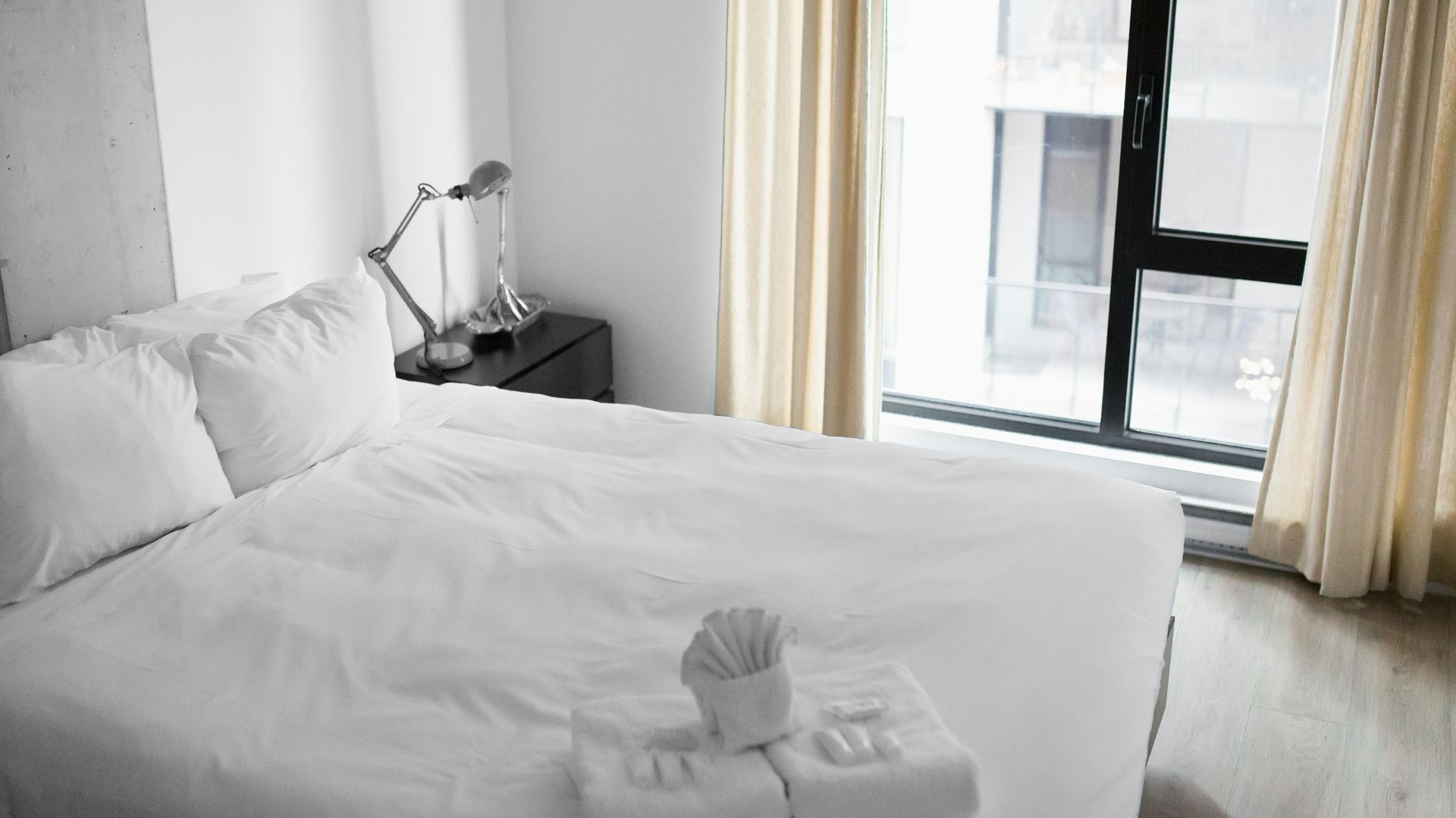 A white bed with folded towels sits in a minimalist bedroom next to a window with beige curtains and a bedside lamp.