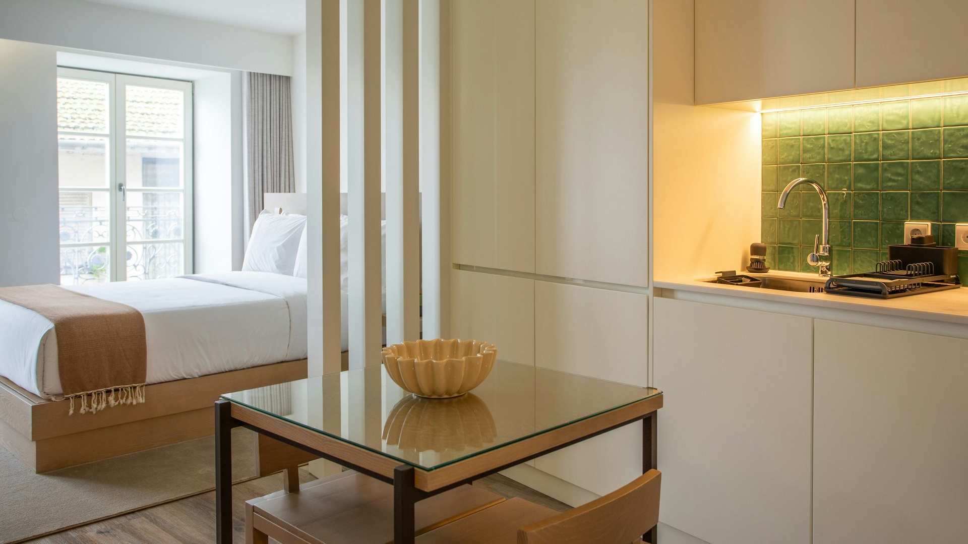 Modern bedroom with a glass table, white bed, and warm-lit kitchenette area