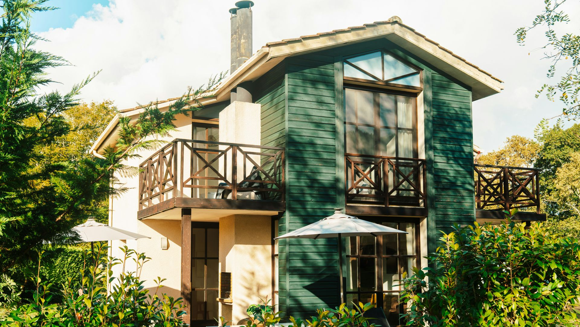 A two-story house with dark green paneling and light beige walls, surrounded by trees with balconies on both levels.