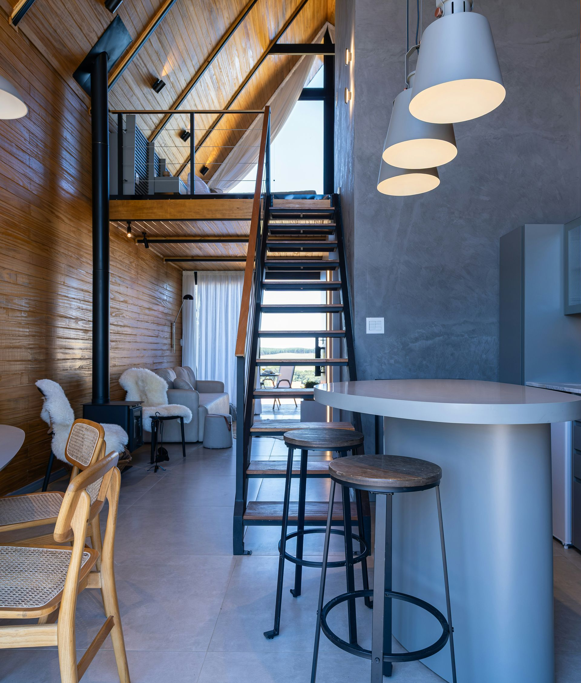 A modern, open-concept living space with a wooden A-frame ceiling, a loft area, a metal staircase, and a breakfast bar.