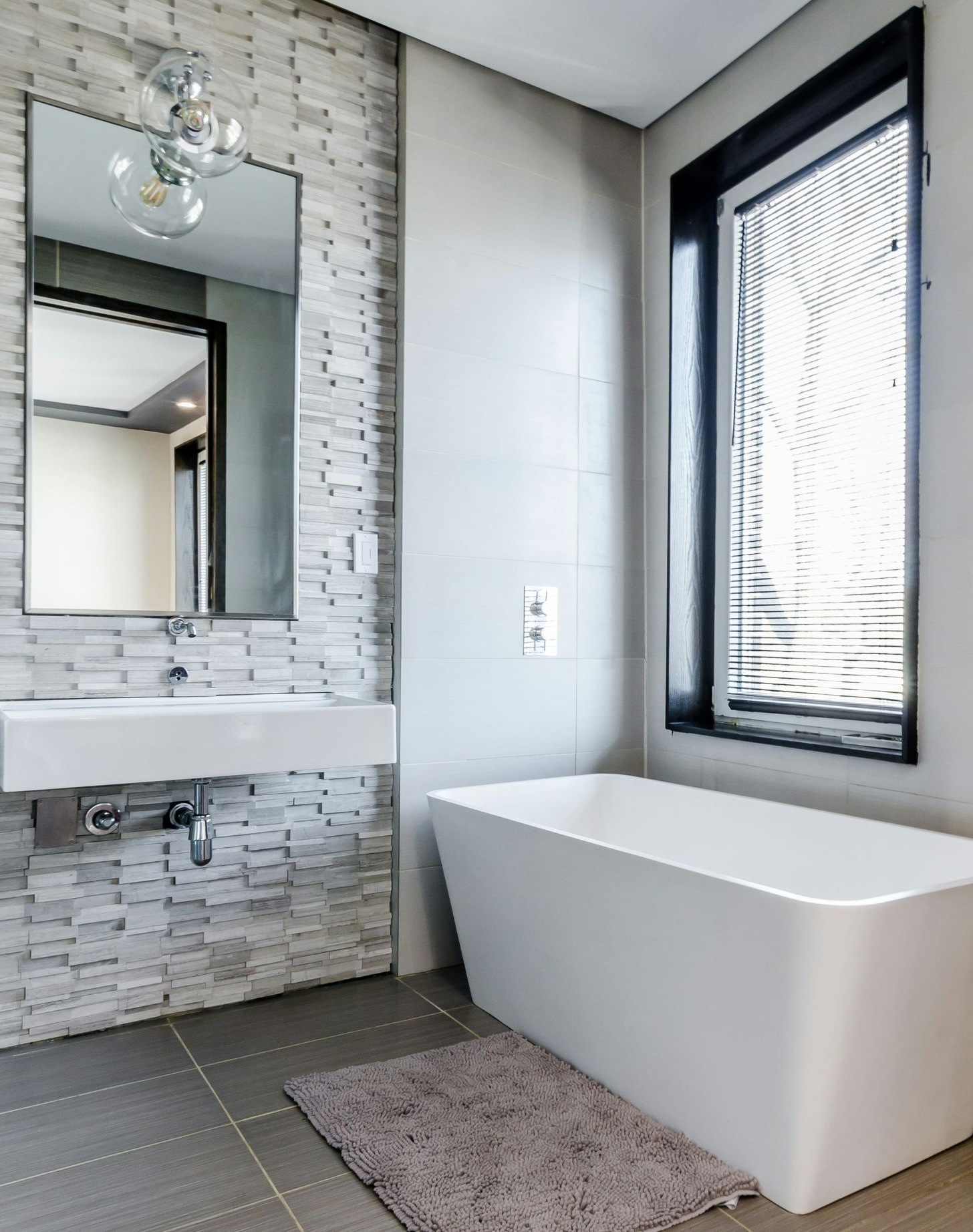 Modern bathroom with freestanding white tub, wall mirror, sink, and large window beside a gray tiled wall.