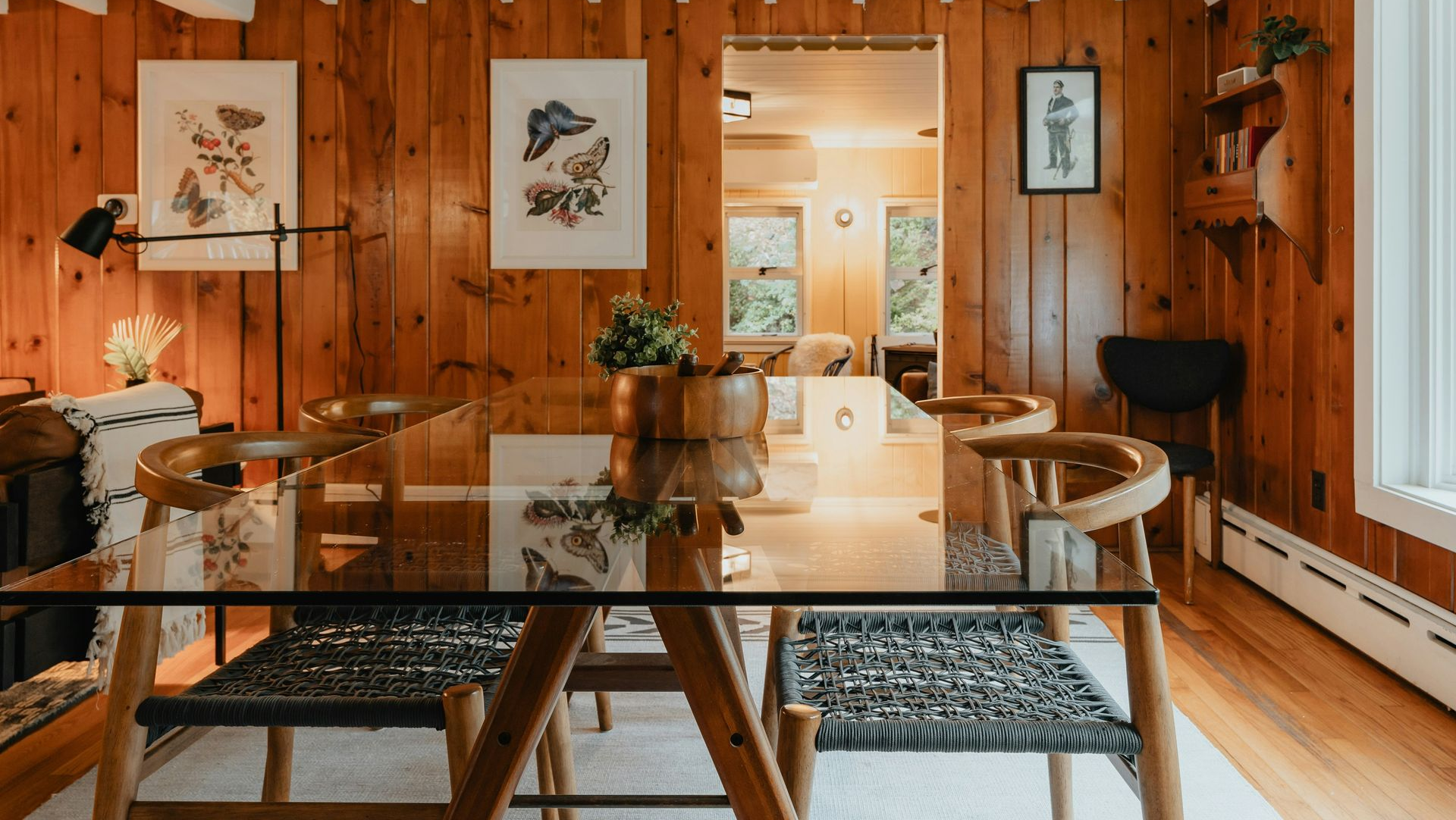 Warm wood-paneled dining room with glass table, woven chairs, patterned rug, and framed art on the walls