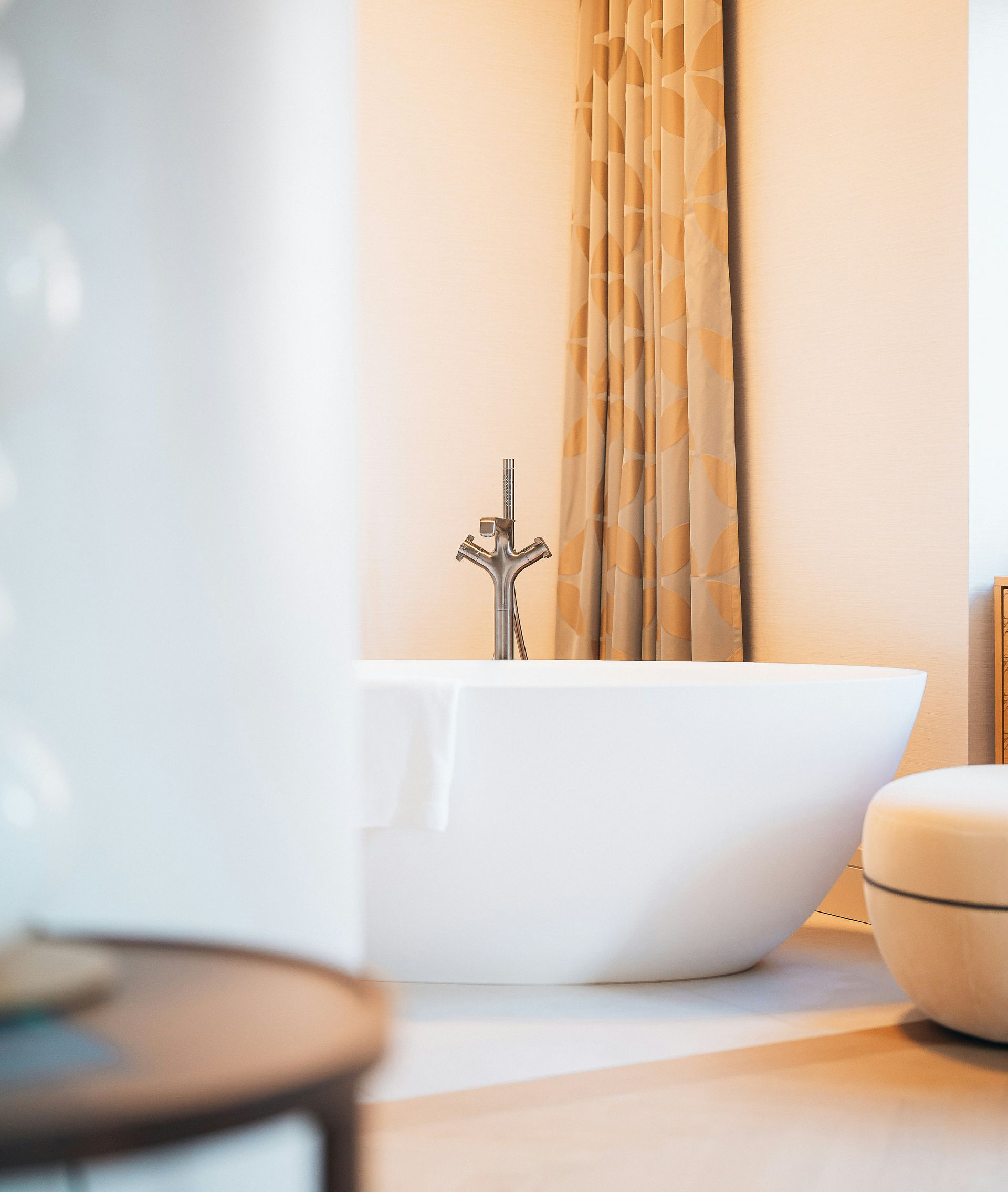 A modern white freestanding bathtub with a silver faucet stands in front of floor-length curtains in a brightly lit room.