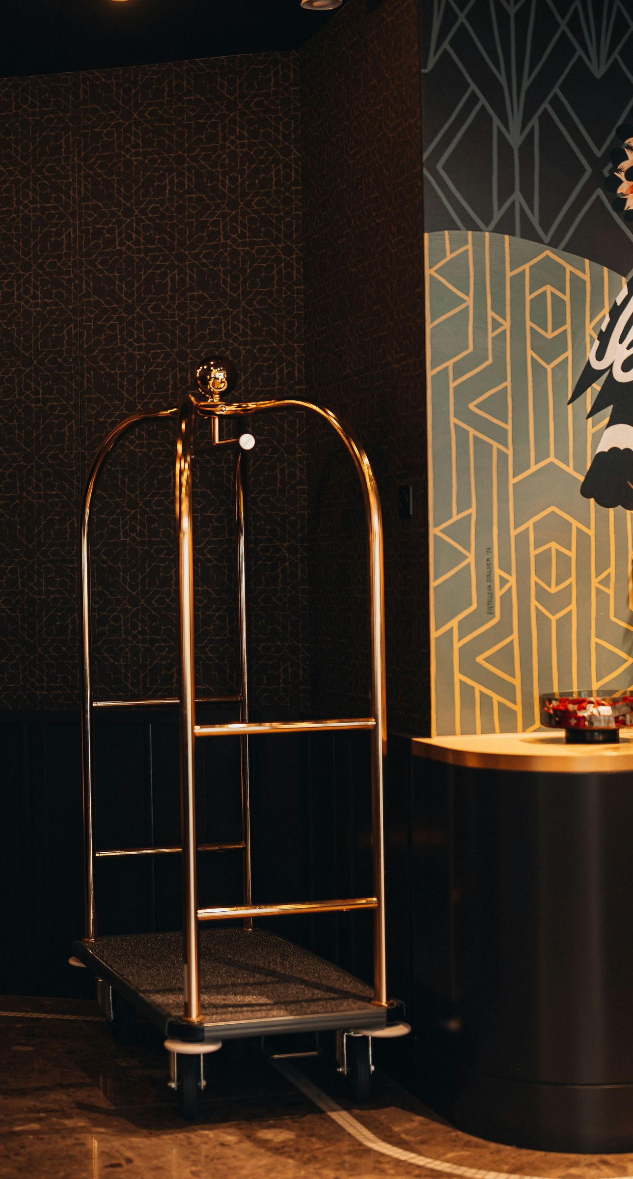 A gold hotel luggage cart stands on a dark floor in a lobby with a decorative geometric patterned wall.